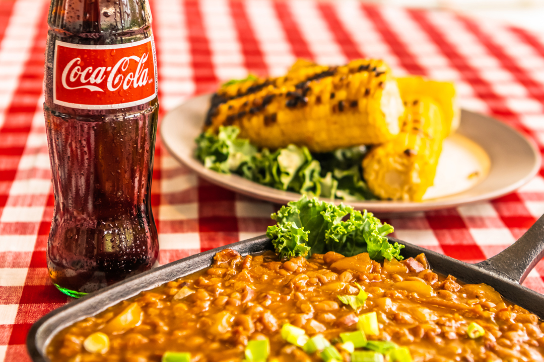 baked beans and glass bottle of coke with stack of corn on the cob in the background