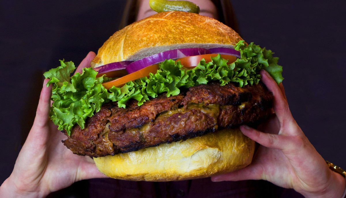 two hands holding up burger on bun with lettuce, tomatoes and onion